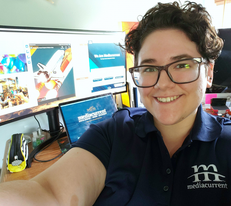 A selfie of a young woman with short curly hair and a navy blue Mediacurrent branded polo. In the background is a laptop and monitor open to DrupalCon, CelebrateDrupal.org, and Mediacurrent.com