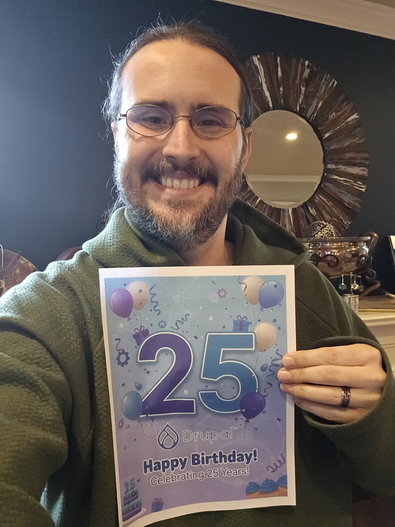 Tim Lehnen (hestenet) holds a sign celebrating Drupal's 25th birthday and smiles.
