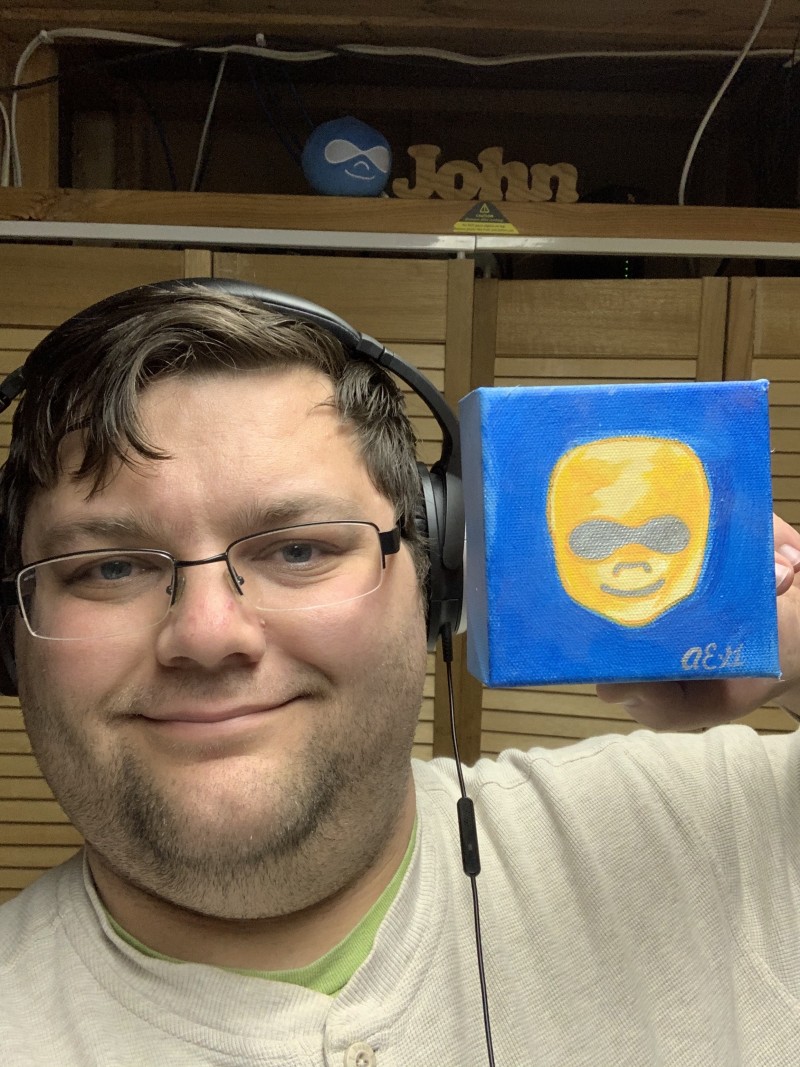 John Rearick holding a small painting of a DrupalCorn logo. A druplicon plush sits in the background