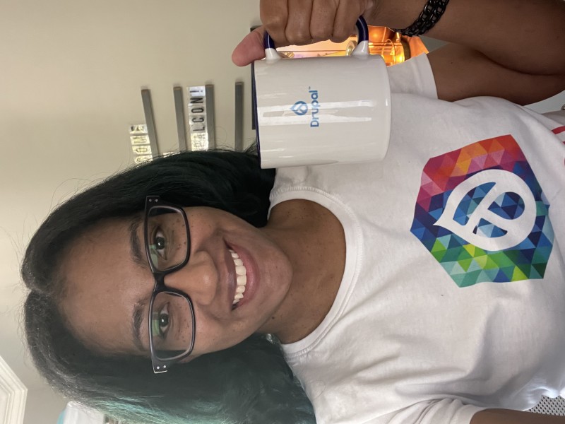 Nicole is wearing a DrupalCon NA shirt, holding a Drupal mug and smiling. 