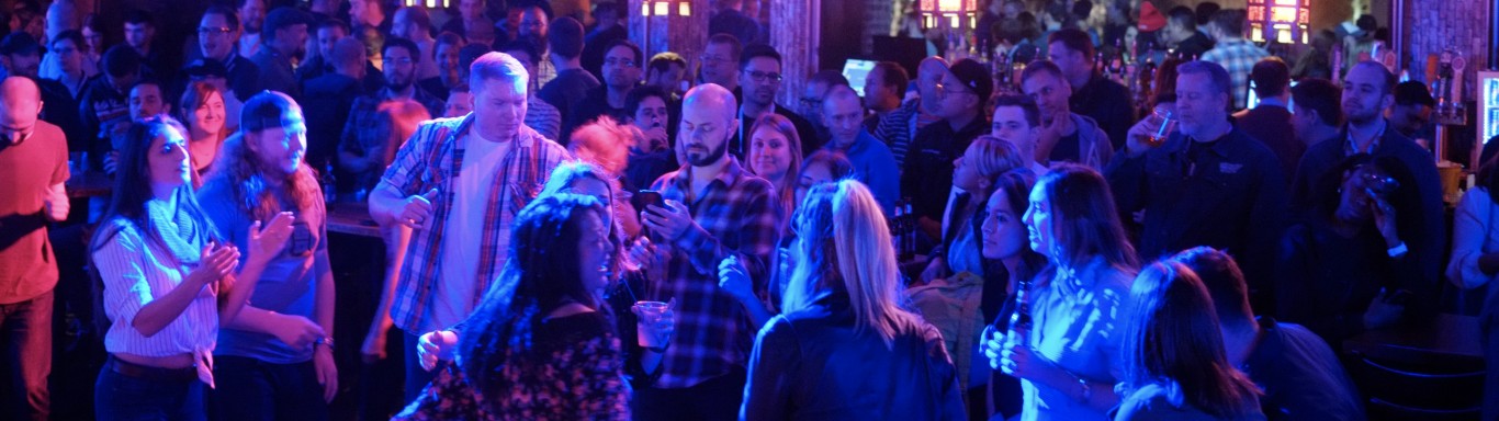 A crowded DrupalCon social event in a dimly lit bar with neon lighting