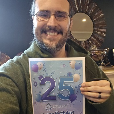 Tim Lehnen (hestenet) holds a sign celebrating Drupal's 25th birthday and smiles.