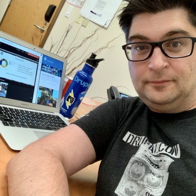 Selfie of a man wearing a DrupalCon shirt in front of a laptop displaying DrupalCon content