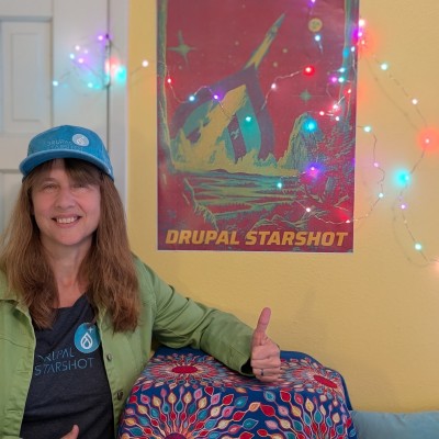 Kristen Pol, a long time Drupal community member, showing off Drupal Starshot swag (tshirt, hat, and poster) in bright colors of teal, red, yellow and more