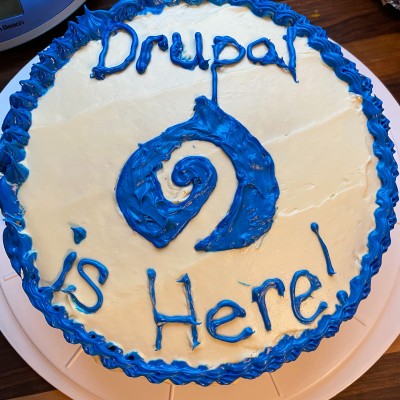 Image of round cake with white frosting—on top in blue icing is the Drupal drop with a 9 inside and text: Drupal Is Here